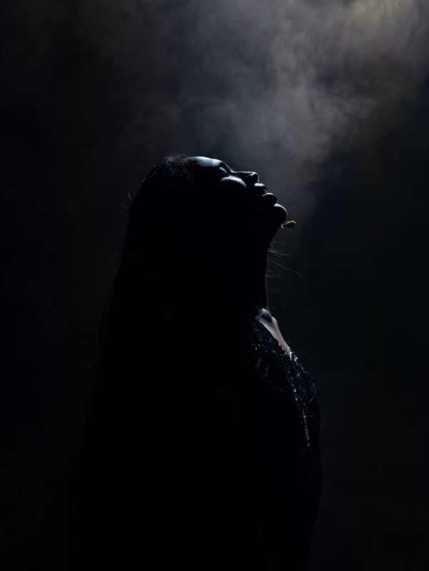 Silhouette of a person with long hair looking upwards in a dark setting, illuminated by subtle lighting from above, with visible smoke in the background.