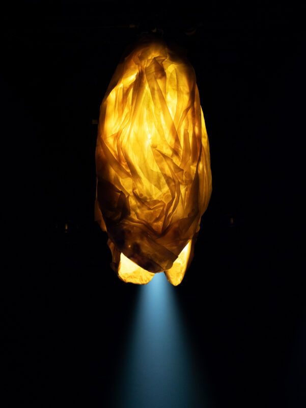 A glowing, crumpled, translucent amber-colored object hangs in darkness with a blue light beam shining from below.