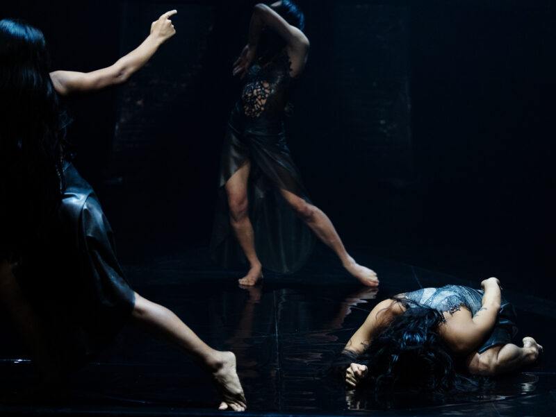 Three dancers perform on a dimly lit stage. One lies on the floor, another stands with an arm covering their face, and a third extends an arm upwards.