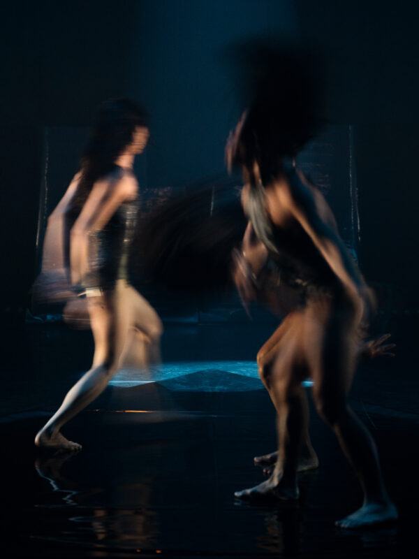 Blurred image of two dancers in motion on a dimly lit stage, creating an abstract effect with their movements.