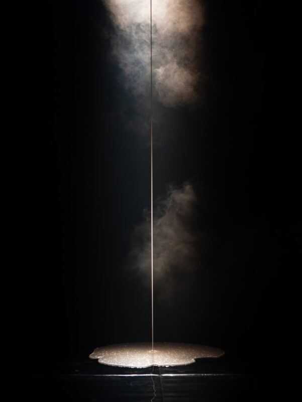 A thin vertical line of light descends through swirling smoke, illuminating a foamy substance on a dark surface below.