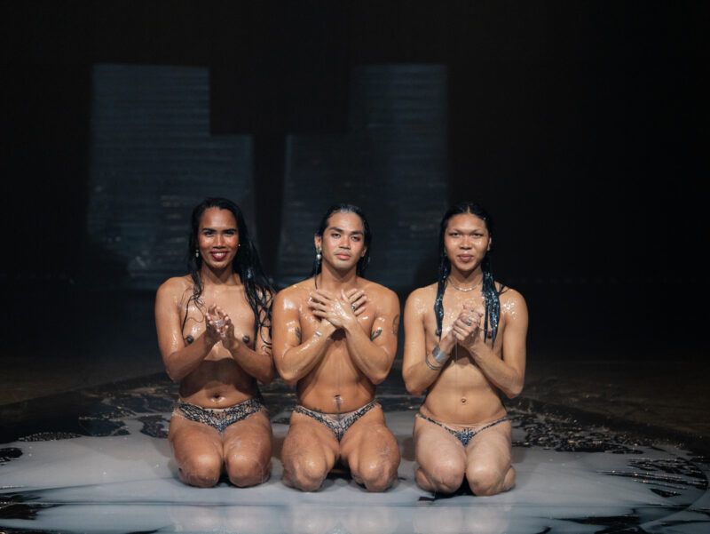 Three people kneel on a wet surface wearing patterned briefs. Their hands are clasped together, and the background is dark.