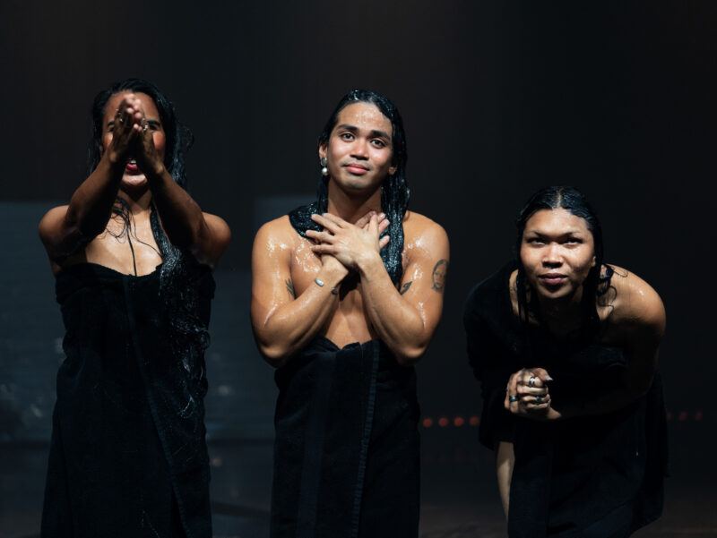 Three people with wet hair, wrapped in black towels, stand together against a dark background.