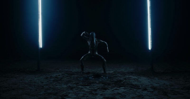 A person stands on their toes with their legs wide, on a dimly lit ground between two bright vertical lights in a dark environment.