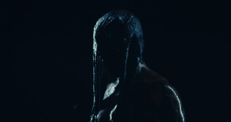 A person covered in a glossy, dark substance stands against a black background, partially illuminated.