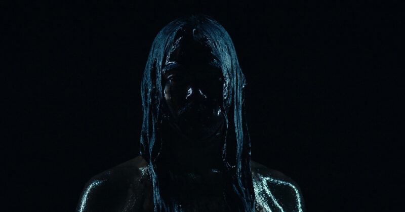 A person stands in low light, covered in a dark, glossy substance, with long hair hanging forward, obscuring most facial features.