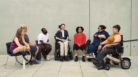 A diverse group of people, some using wheelchairs, sit in a circle and engage in conversation against a plain concrete wall.