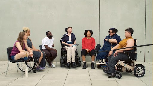 A diverse group of people, some using wheelchairs, sit in a circle and engage in conversation against a plain concrete wall.