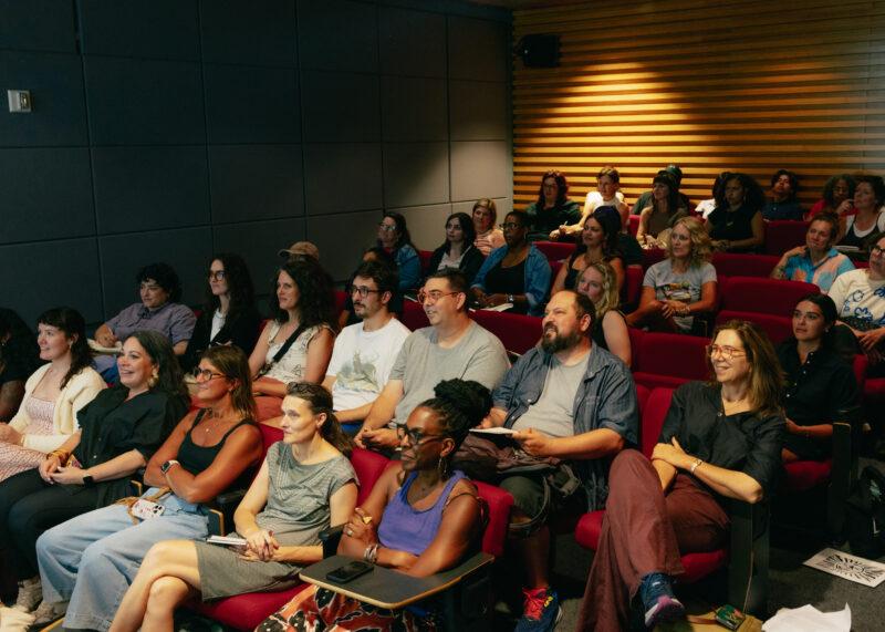 A group of adults sits in red theater seats, attentively watching a presentation in a modern lecture room.