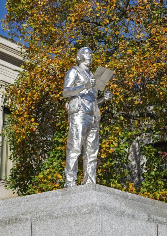 A silver statue of a standing person holding a large piece of paper, set on a stone pedestal, with autumn trees and a building in the background.