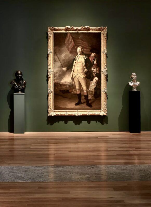 A large framed portrait of a man in historical attire hangs on a dark green wall, flanked by two bust sculptures on pedestals in a museum gallery.