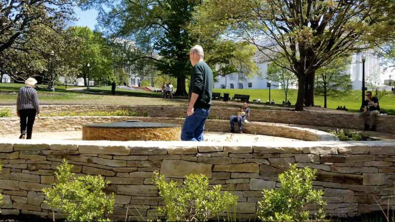 Several people walk and sit near a circular stone structure in a park with trees and greenery on a sunny day.