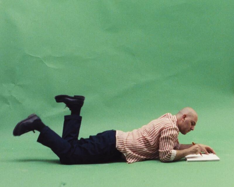 Hadi Falapishi lies on his stomach on a green background, writing or drawing in an open notebook.