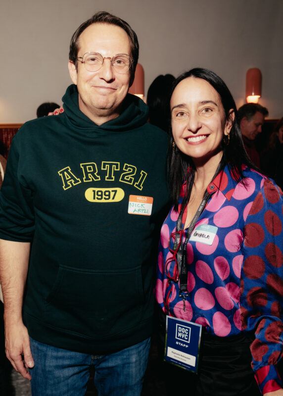 Two people stand together indoors at an event, both smiling at the camera. One wears a dark hoodie with "ART21 1997" and the other wears a pink-patterned shirt with a staff badge.