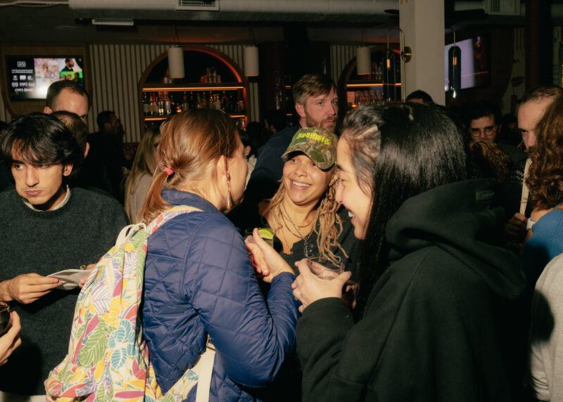 A group of people socializes at a lively indoor bar or restaurant, holding drinks and chatting in a warmly lit setting.