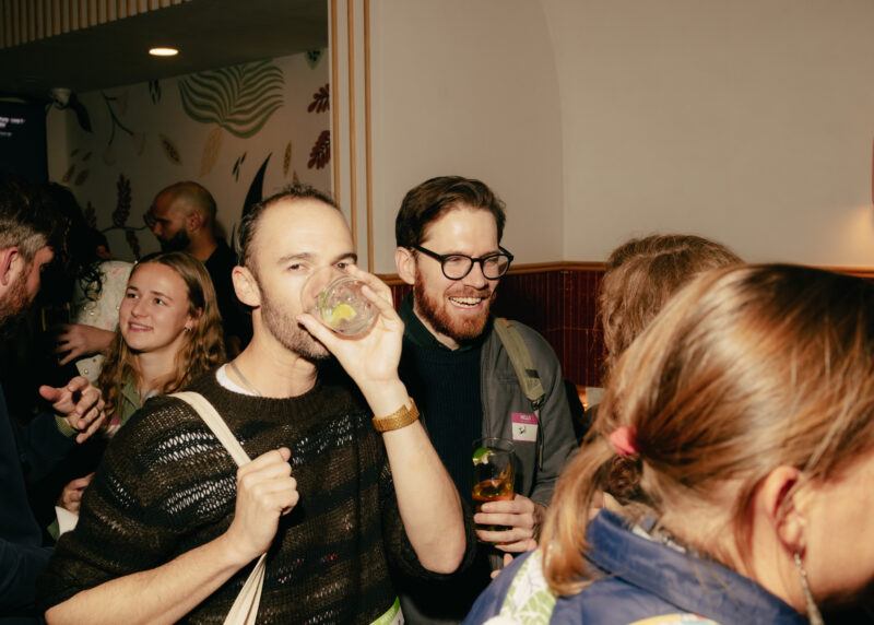A group of people stand closely together at a social gathering; one man drinks from a glass while others talk and smile.