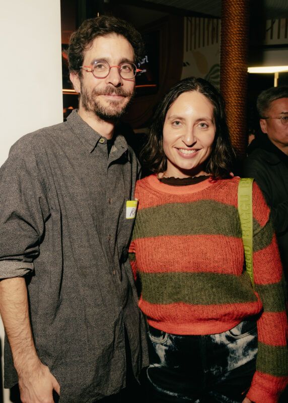 Two people pose together indoors at a social event; one wears glasses and a gray shirt, the other wears a red and green striped sweater.