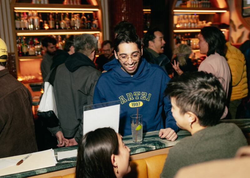 A man in a blue Art21 hoodie stands at a bar counter, smiling and talking to two people, with a crowd and shelves of bottles in the background.