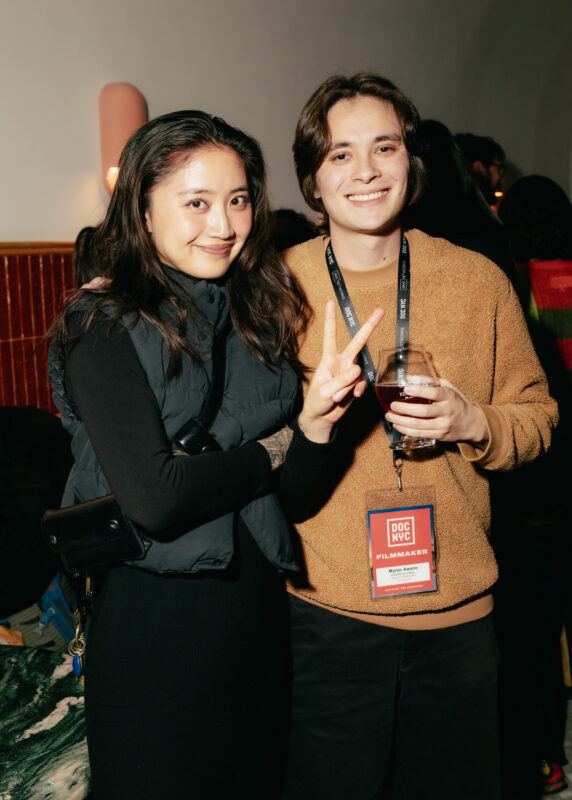 Two people pose together at an event; one holds a drink and wears a "Filmmaker" badge, while the other smiles and holds up a peace sign.