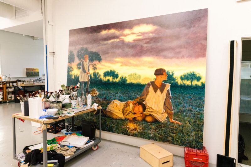A large, realistic painting depicting three people in a field is propped against a wall in an art studio, with art supplies and a table in the foreground.