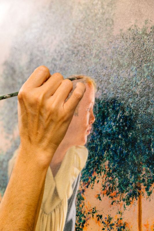 A close-up of a hand holding a paintbrush, painting a realistic portrait of a person and trees on a canvas.