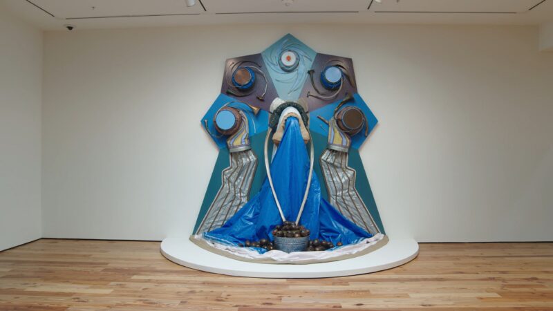 Sculptural art installation featuring flowing blue fabrics, circular elements, and metallic accents displayed against a white gallery wall on a round platform.