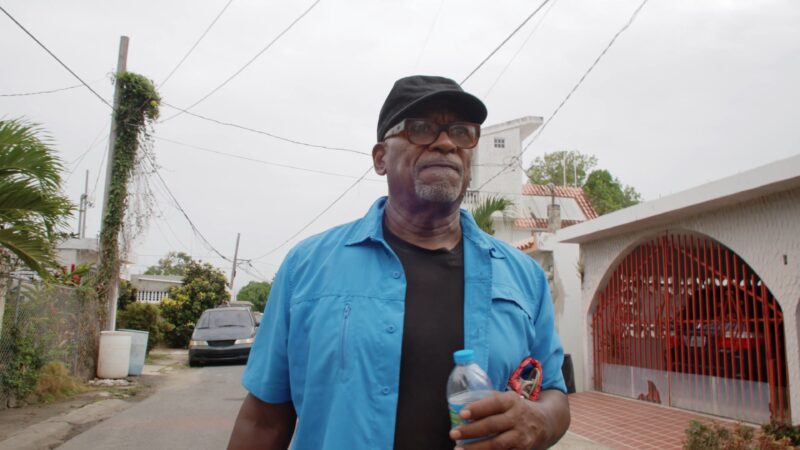 Daniel Lind-Ramos wearing a blue shirt, black cap, and glasses holds a water bottle while standing on a residential street with houses, power lines, and a parked car visible.
