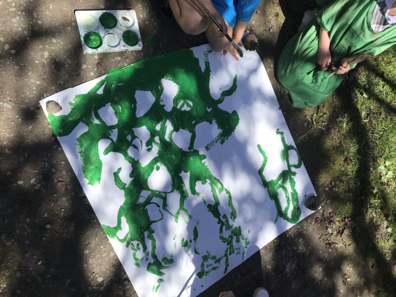 Aerial view of children painting abstract green shapes on a large white sheet outdoors, with paint palette and brushes visible.