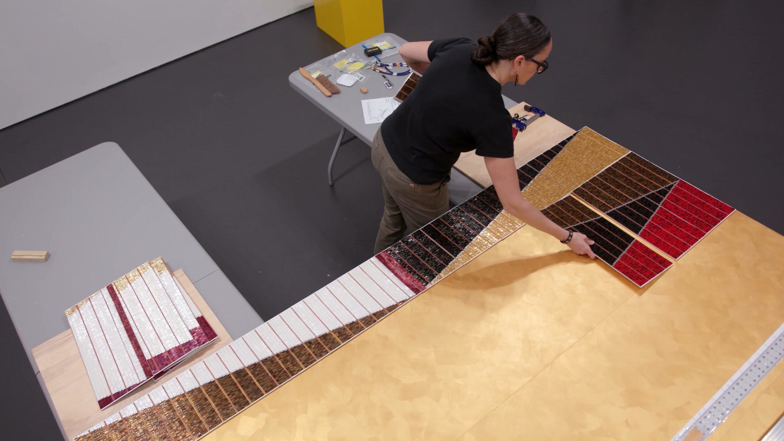 Dyani White Hawk applies beaded panels to a large flat surface, with tools and supplies on a nearby table in a workspace.