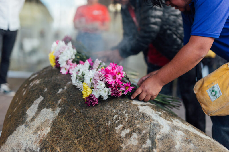 Person in a blue shirt places colorful flowers on a large stone, with other people and a blurred background visible.