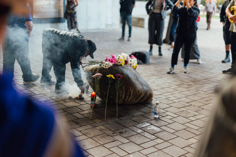 People gather around a memorial with a large stone adorned with flowers, candles, and a water bottle on the ground. Smoke rises as one person leans over the stone.