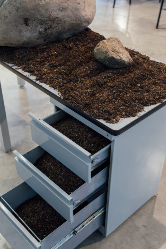 A metal desk with three drawers partially open, filled with soil. Dirt covers the desk surface, with two large rocks placed on top.