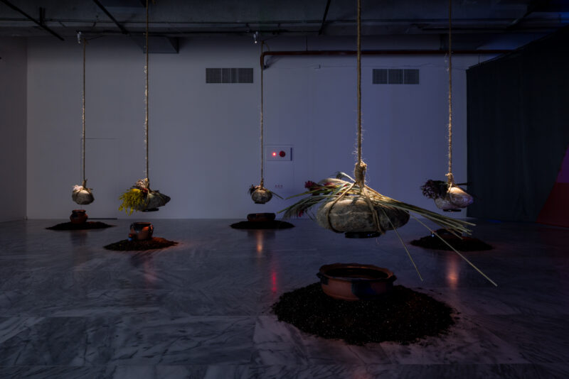 Dimly lit installation with five large stones wrapped in rope, suspended above clay pots placed on circular soil piles on a marble floor.