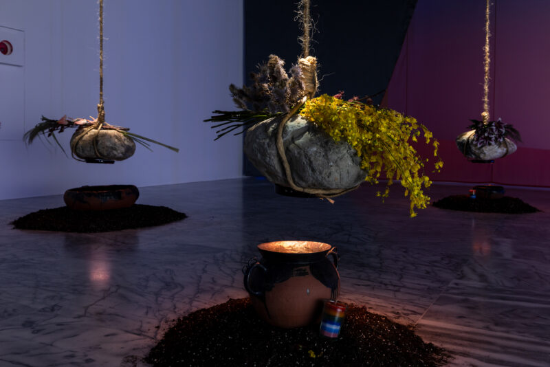 Three large stones wrapped with rope, decorated with flowers, are suspended above clay pots filled with soil on a marble floor in a dimly lit room.