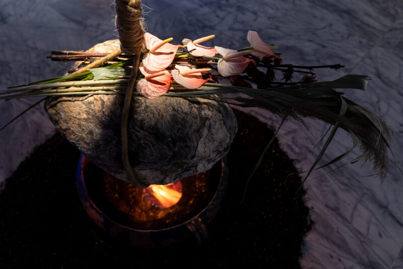 A bundle of pink flowers and green foliage tied to a large stone hovers above an open flame, with the setup placed on a marble floor.