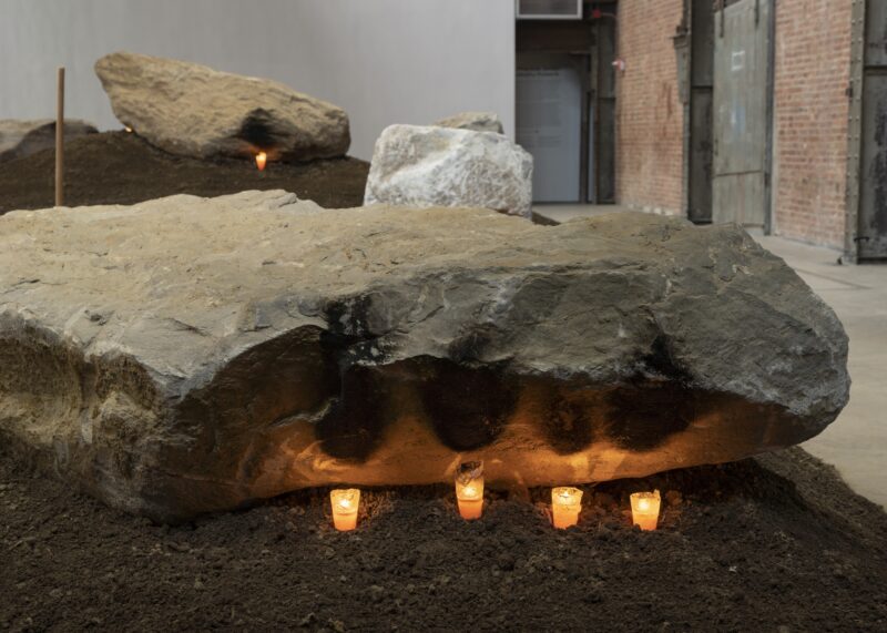 A large stone rests on dirt indoors, with five lit candles placed beneath its edge; other rocks and dirt mounds are visible in the background.