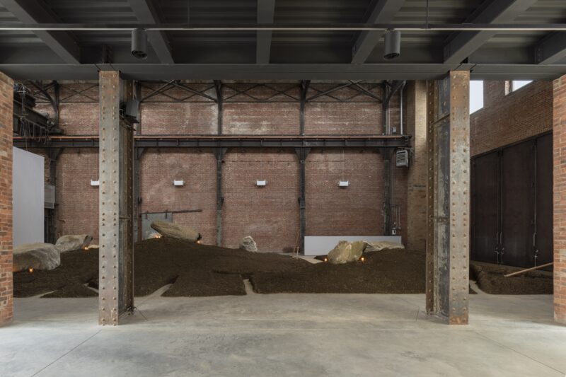 An indoor installation featuring a mound of soil with large rocks, set against exposed brick walls and industrial steel beams in a spacious gallery.