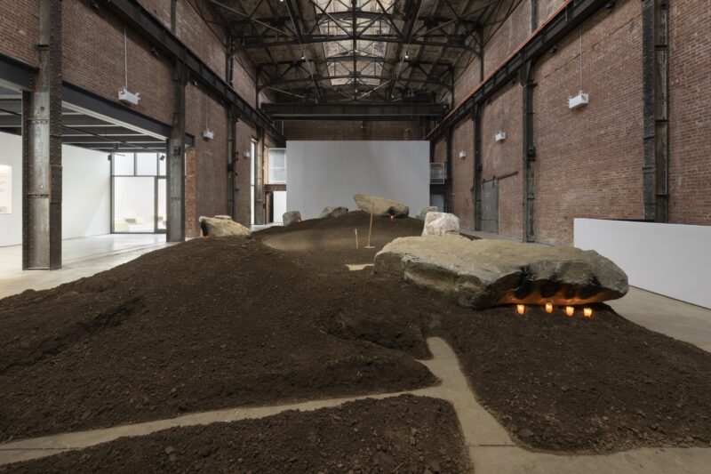 Large indoor art installation featuring mounds of soil and several large rocks, some illuminated from underneath, set in an industrial-style brick building.