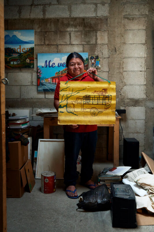 Edgar Calel stands in a room with art supplies, holding a yellow painting of a bus. Other paintings and various items are visible around him against a bare concrete wall.