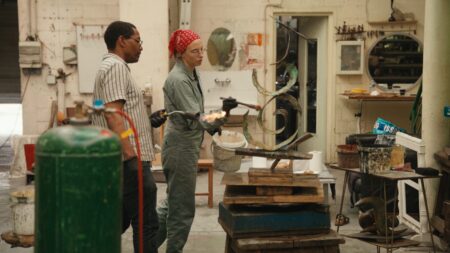 Two people in a workshop use torches to work on a metal sculpture; tools, gas tanks, and art supplies are visible around them.
