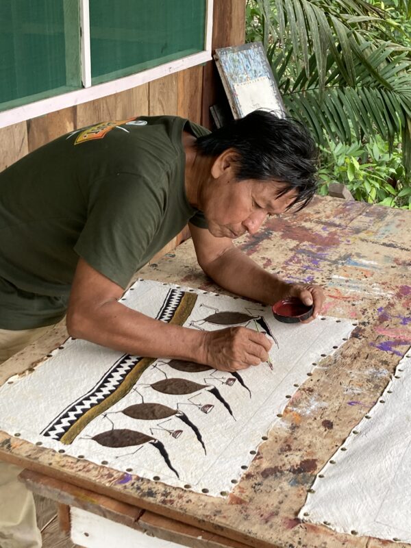 Santiago Yahuarcani carefully paints on a textile laid out on a wooden table, surrounded by greenery and natural light.