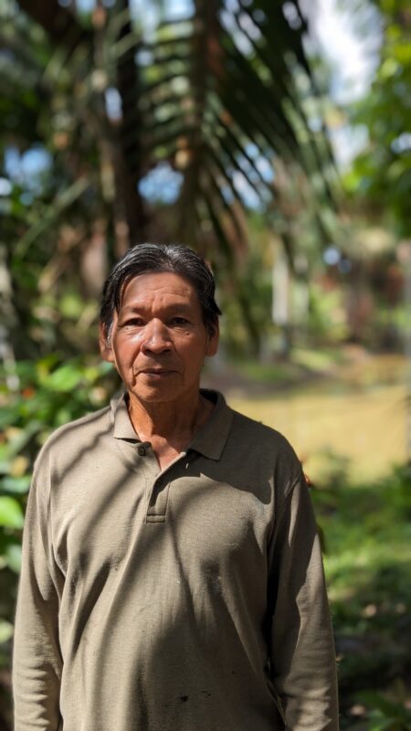 Santiago Yahuarcani with dark hair stands outdoors in a garden, wearing a tan collared shirt. Sunlight and shadows fall across his face and body, with green foliage in the background.
