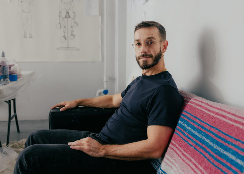 Carlos Motta with glasses and a beard sits on a couch with a striped pink and blue blanket, in a room with sketch drawings on the wall.