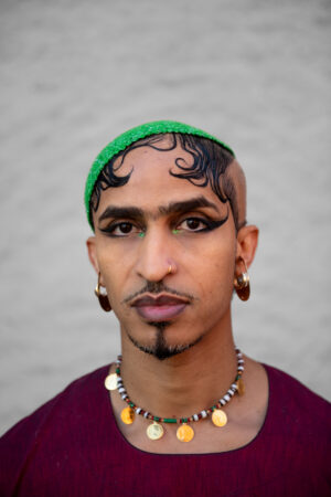 Ahmed Umar with decorative facial hair and bold eye makeup wears a green headpiece, gold hoop earrings, a coin necklace, and a maroon top, standing against a plain background.