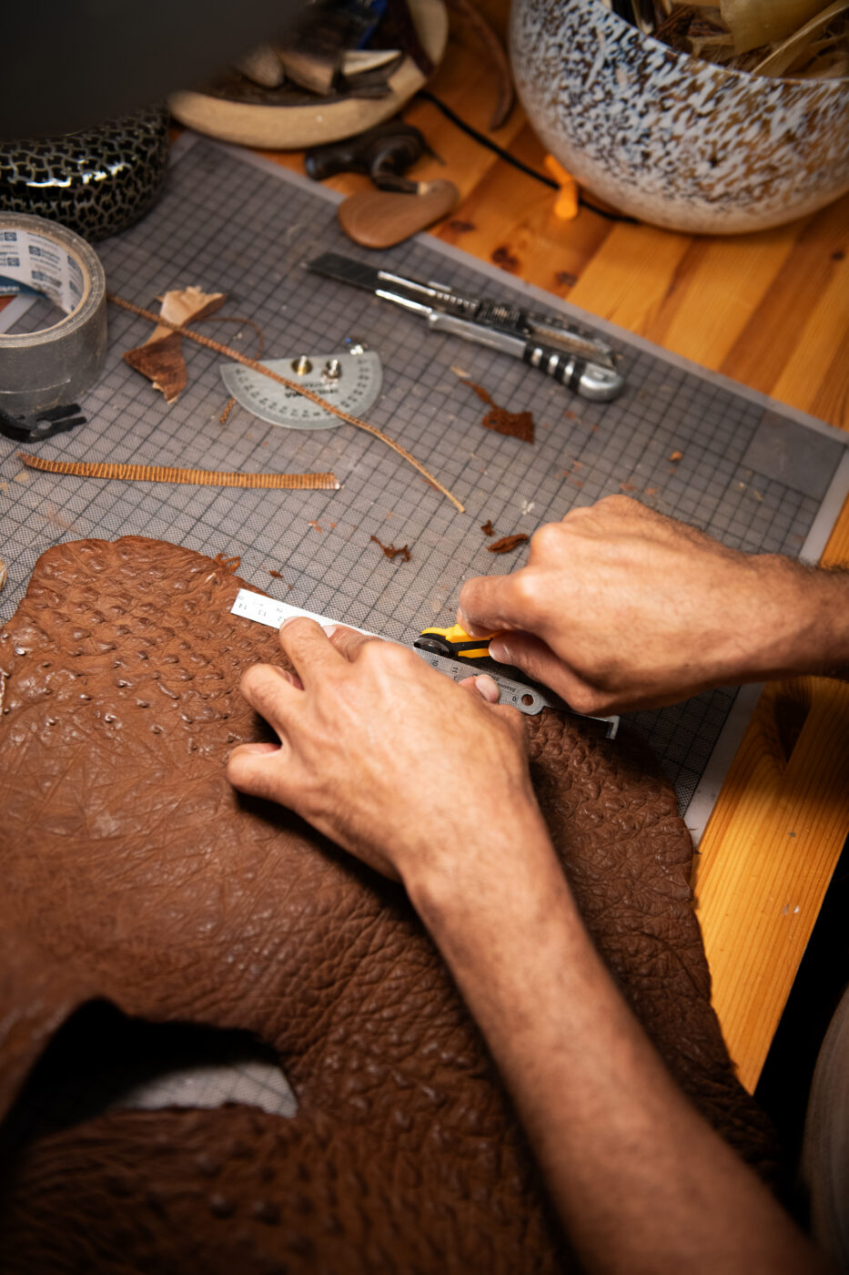 A person cuts a piece of textured brown leather on a worktable with various crafting tools, a ruler, and measuring grid visible.