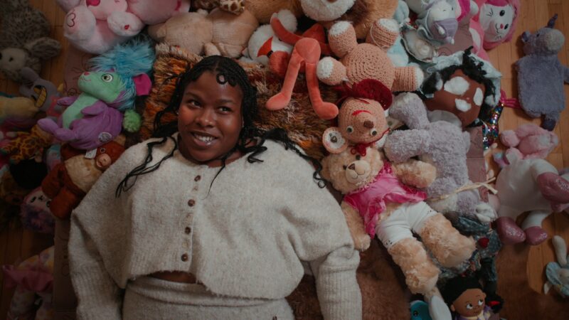 Precious Okoyomon with braided hair and a light sweater lies on a textured rug, smiling, surrounded by colorful stuffed toys.