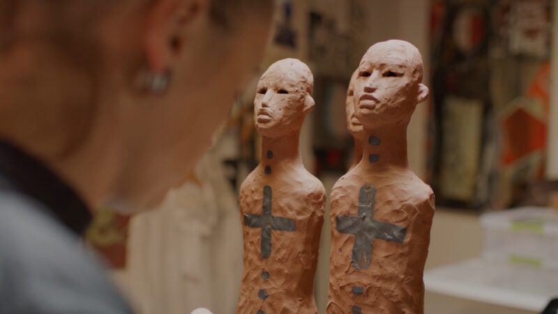 A person observes two clay sculptures of human figures with dark cross marks and dots painted on their torsos.