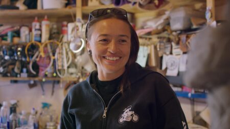 Rose B. Simpson with long brown hair and sunglasses on their head smiles in a workshop filled with tools and equipment.