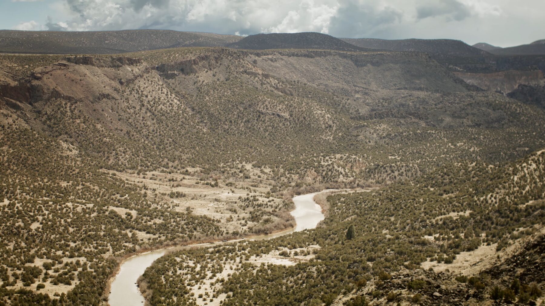 A winding river flows through a dry, hilly landscape covered with sparse vegetation, with distant mountains under a cloudy sky.