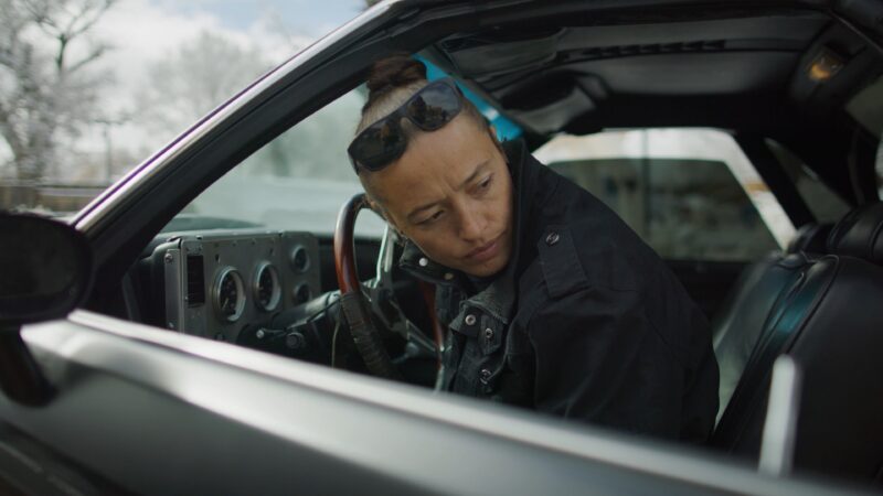 Rose B. Simpson sitting in the driver's seat of a car, looking back over their shoulder with sunglasses resting on their head.